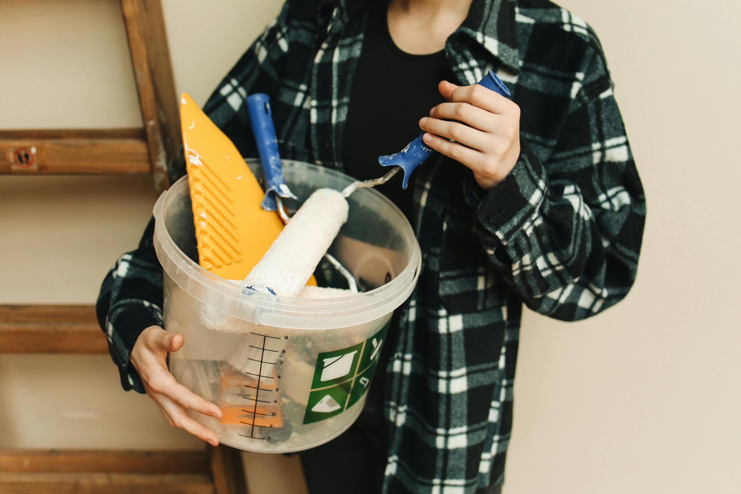 woman in a plaid shirt holding a bucket of wall repair tools next to a ladder